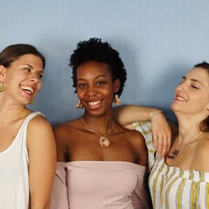 May include: Three women are smiling and laughing together. The woman in the middle is wearing a pink off-the-shoulder top and a brown pendant necklace. The woman on the left is wearing a white tank top and green earrings. The woman on the right is wearing a white and yellow striped top and brown earrings.