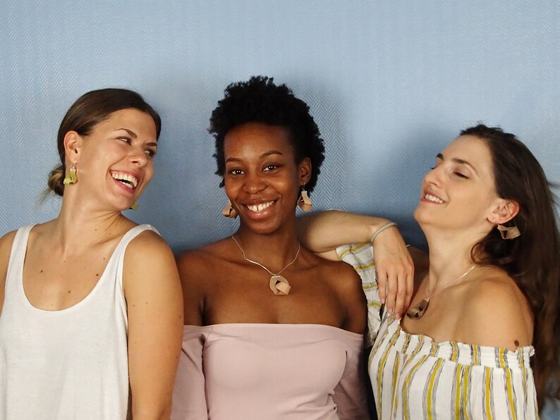 May include: Three women are smiling and laughing together. The woman in the middle is wearing a pink off-the-shoulder top and a brown pendant necklace. The woman on the left is wearing a white tank top and green earrings. The woman on the right is wearing a white and yellow striped top and brown earrings.