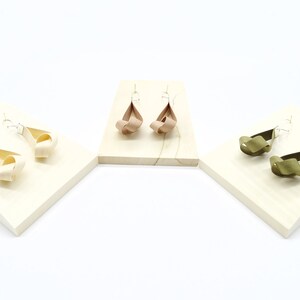 May include: Three pairs of earrings on display stands. The earrings are made of fabric and are tied in a knot. The earrings are in three colors: beige, brown, and green.