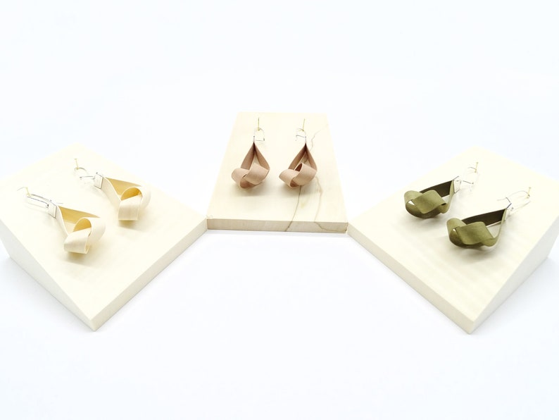 May include: Three pairs of earrings on display stands. The earrings are made of fabric and are tied in a knot. The earrings are in three colors: beige, brown, and green.