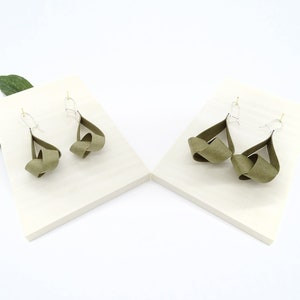 May include: A pair of olive green knotted dangle earrings with silver hooks. The earrings are displayed on two white surfaces.