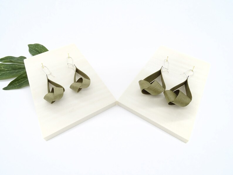 May include: A pair of olive green knotted dangle earrings with silver hooks. The earrings are displayed on two white surfaces.