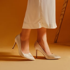 May include: A pair of ivory satin pumps with stiletto heels. The shoes are on a light brown surface.