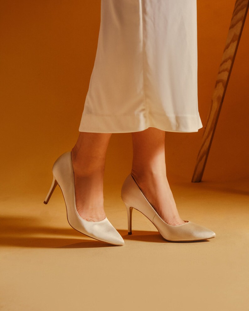 May include: A pair of ivory satin pumps with stiletto heels. The shoes are on a light brown surface.