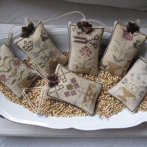 May include: Six small, rectangular, beige fabric pillows with embroidered designs. The pillows are arranged in a white oval dish filled with beige and brown birdseed.