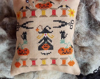 Op de afbeelding: Een beige stoffen naaldkussen met een kruissteekontwerp met een heks, pompoenen en spoelen garen. Het naaldkussen heeft een zwarte kwast.