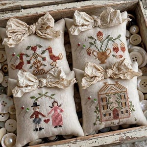 Puede incluir: Cuatro pequeños alfileteros cuadrados de tela beige, cada uno con un lazo de tela arrugada. Los cojines presentan diseños en punto de cruz de dos pájaros, un jarrón floral, una pareja y una casa, colocados entre botones de color blanco.