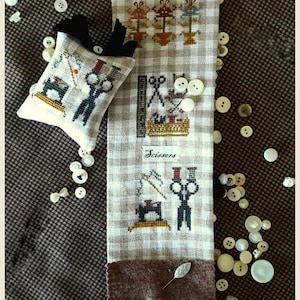 May include: A white and brown fabric sampler with cross-stitch designs of sewing tools, including scissors, a needle, and a sewing machine. The sampler is surrounded by white buttons and a brown fabric pincushion with a cross-stitch design of sewing tools.