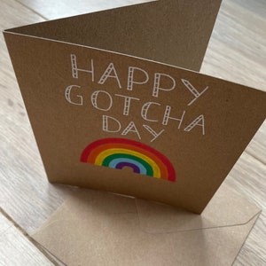 May include: A brown card with the text "Happy Gotcha Day" in white outlined letters. A rainbow with red, orange, yellow, green, blue, indigo, and violet stripes is below the text.