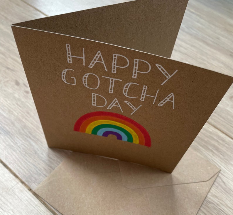 May include: A brown card with the text "Happy Gotcha Day" in white outlined letters. A rainbow with red, orange, yellow, green, blue, indigo, and violet stripes is below the text.