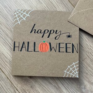 May include: A square, brown card with the words "happy HALLOWEEN" in black script and block letters. An orange pumpkin is centered below the word "happy". White spiderwebs are in the corners, and a small black spider is above the word "Halloween".