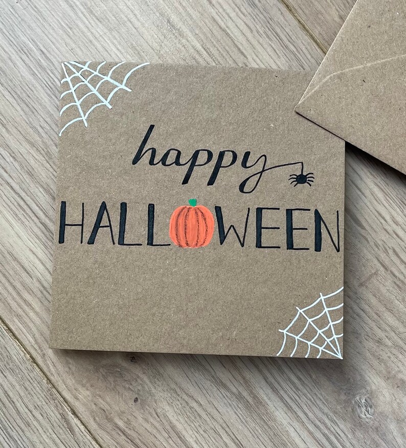 May include: A square, brown card with the words "happy HALLOWEEN" in black script and block letters. An orange pumpkin is centered below the word "happy". White spiderwebs are in the corners, and a small black spider is above the word "Halloween".