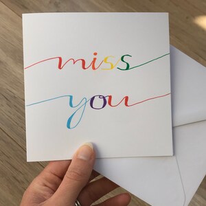 May include: A white card with the words "miss you" written in rainbow colors. The card is held in a hand.