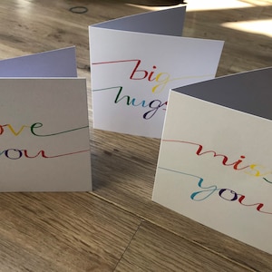 May include: Three white greeting cards with rainbow colored lettering. The cards say "love you", "big hugs", and "miss you".