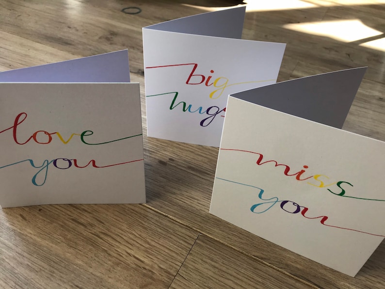 May include: Three white greeting cards with rainbow colored lettering. The cards say "love you", "big hugs", and "miss you".