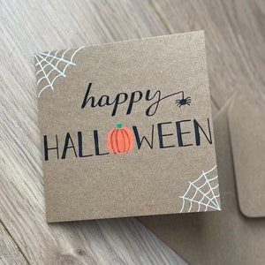 May include: A square, brown Halloween card with the words "happy HALLOWEEN" in black script and block letters. An orange pumpkin graphic is between the words. White spiderwebs are in the corners, and a black spider is above the word "HALLOWEEN".