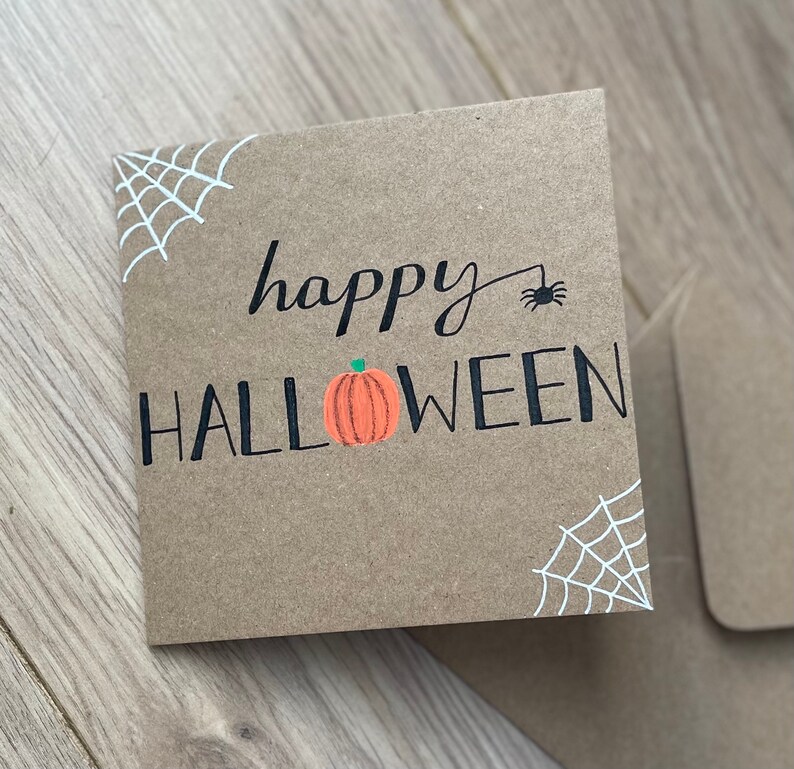 May include: A square, brown Halloween card with the words "happy HALLOWEEN" in black script and block letters. An orange pumpkin graphic is between the words. White spiderwebs are in the corners, and a black spider is above the word "HALLOWEEN".