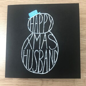 May include: A black greeting card with white lettering that reads "HAPPY XMAS HUSBAND". The words are arranged in the shape of a snowman, with a small blue hat on top. A festive holiday card.