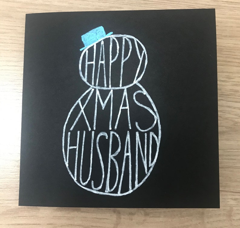 May include: A black greeting card with white lettering that reads "HAPPY XMAS HUSBAND". The words are arranged in the shape of a snowman, with a small blue hat on top. A festive holiday card.