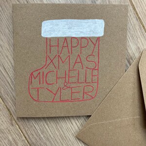 May include: A square, kraft paper Christmas card with a red stocking design. The stocking contains the words "HAPPY XMAS MICHELLE & TYLER" in red. The top of the stocking is white. A matching envelope is next to the card.