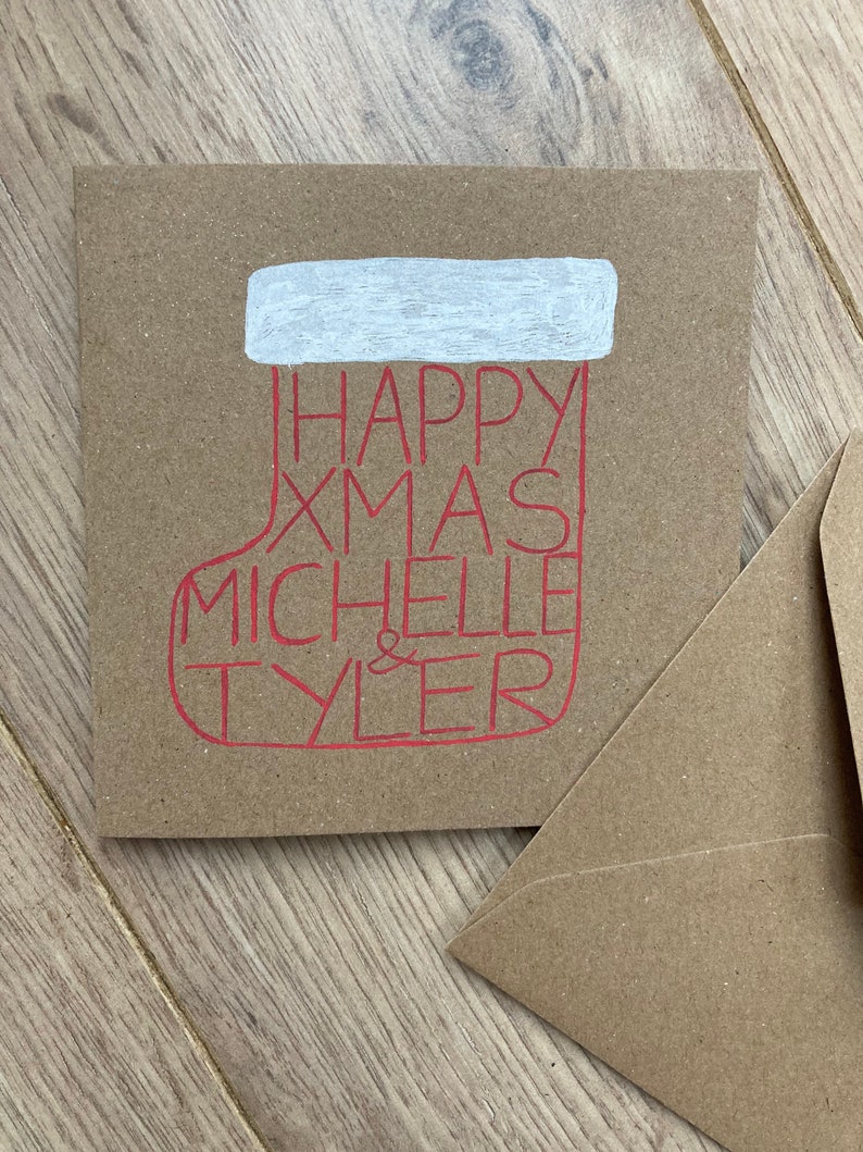 May include: A square, kraft paper Christmas card with a red stocking design. The stocking contains the words "HAPPY XMAS MICHELLE & TYLER" in red. The top of the stocking is white. A matching envelope is next to the card.