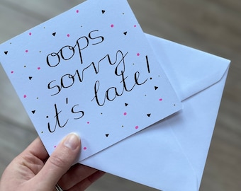May include: A white card with pink and black confetti printed on it. The card has black calligraphy that reads "oops sorry it's late!"