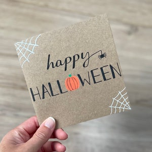 May include: A square, brown card with the words "happy HALLOWEEN" in black script and block letters. An orange pumpkin with a green stem is between the words. White spiderwebs are in the corners, and a black spider is above the word "HALLOWEEN".