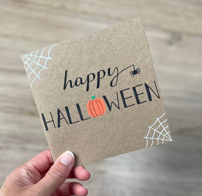 May include: A square, brown card with the words "happy HALLOWEEN" in black script and block letters. An orange pumpkin with a green stem is between the words. White spiderwebs are in the corners, and a black spider is above the word "HALLOWEEN".