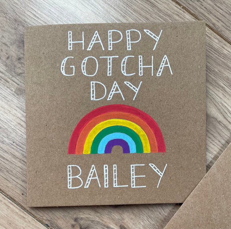 May include: A brown card with the text "Happy Gotcha Day Bailey" in white. A rainbow is drawn below the text.
