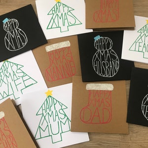 May include: A collection of Christmas greeting cards with various designs. Some cards feature a snowman or Christmas tree shape, while others are shaped like a stocking. The words "Happy Xmas" are incorporated into the designs, with additional words like "Teacher", "Husband", and "Dad".