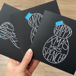May include: Three black Christmas cards with a snowman design. The snowman is drawn in white with the words "HAPPY XMAS MRS SPENCER" inside. The snowman has a blue hat. The cards are held in a hand.
