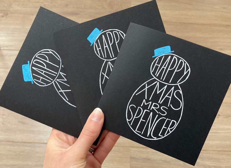 May include: Three black Christmas cards with a snowman design. The snowman is drawn in white with the words "HAPPY XMAS MRS SPENCER" inside. The snowman has a blue hat. The cards are held in a hand.
