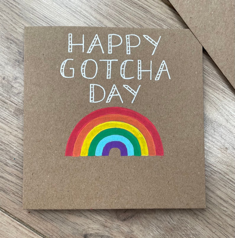 May include: A brown card with the text "Happy Gotcha Day" in white lettering. A rainbow is drawn below the text.