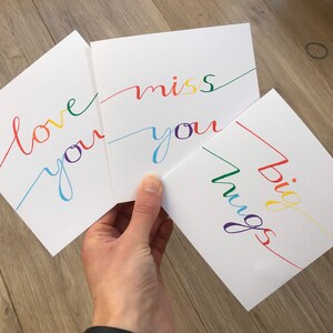 May include: Three white cards with colorful handwritten messages. The first card says "love you" in rainbow colors. The second card says "miss you" in rainbow colors. The third card says "big hugs" in rainbow colors.