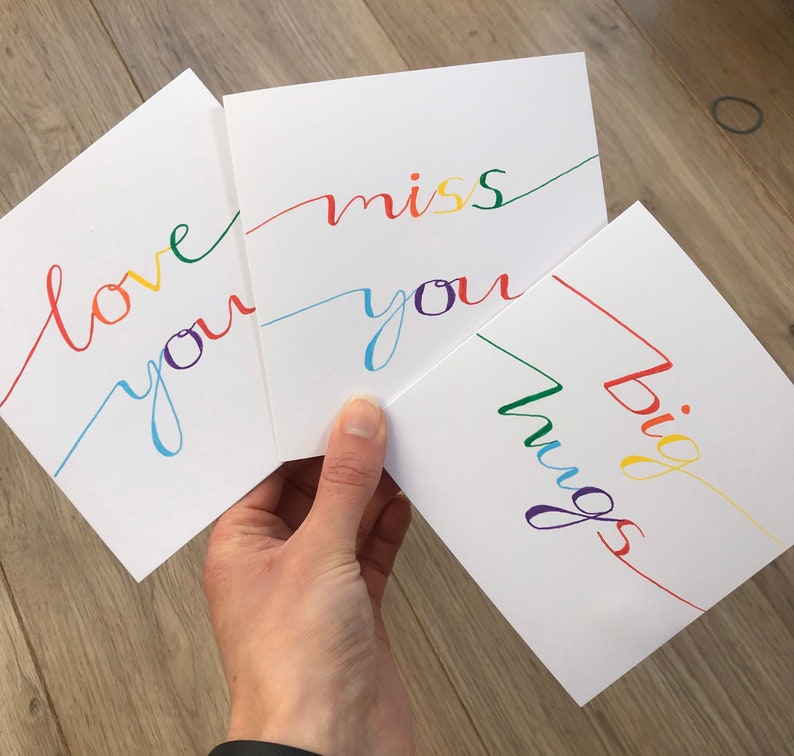 May include: Three white cards with colorful handwritten messages. The first card says "love you" in rainbow colors. The second card says "miss you" in rainbow colors. The third card says "big hugs" in rainbow colors.