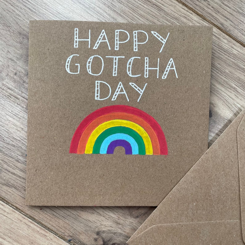 May include: A brown card with the text "Happy Gotcha Day" in white lettering. A rainbow with red, orange, yellow, green, blue, indigo, and violet stripes is drawn below the text.