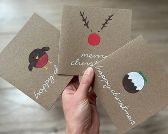 May include: Three square Christmas cards with hand-drawn illustrations. One card features a robin, another a reindeer, and the third a Christmas pudding. Each card has the words "happy christmas" or "merry christmas" in white script.