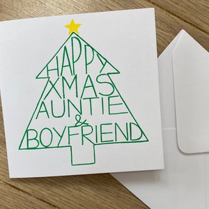 May include: A white Christmas card with a green Christmas tree design. The tree is filled with the words "HAPPY XMAS AUNTIE & BOYFRIEND". A yellow star tops the tree. A white envelope is next to the card.