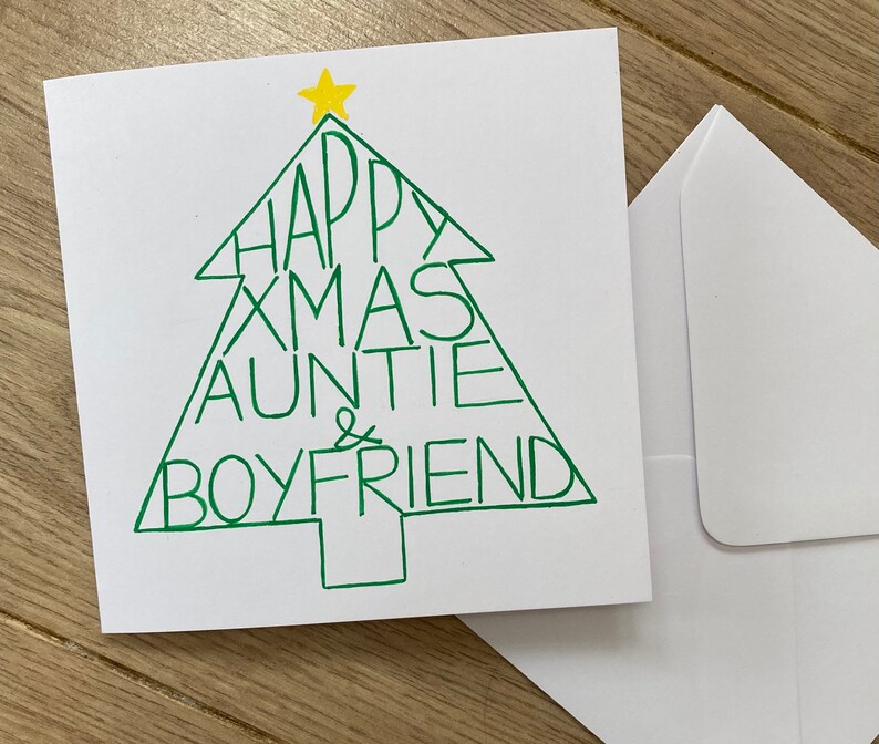 May include: A white Christmas card with a green Christmas tree design. The tree is filled with the words "HAPPY XMAS AUNTIE & BOYFRIEND". A yellow star tops the tree. A white envelope is next to the card.