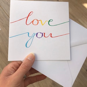 May include: A white card with the words "love you" written in rainbow colors. The card is folded in half and held in a hand.