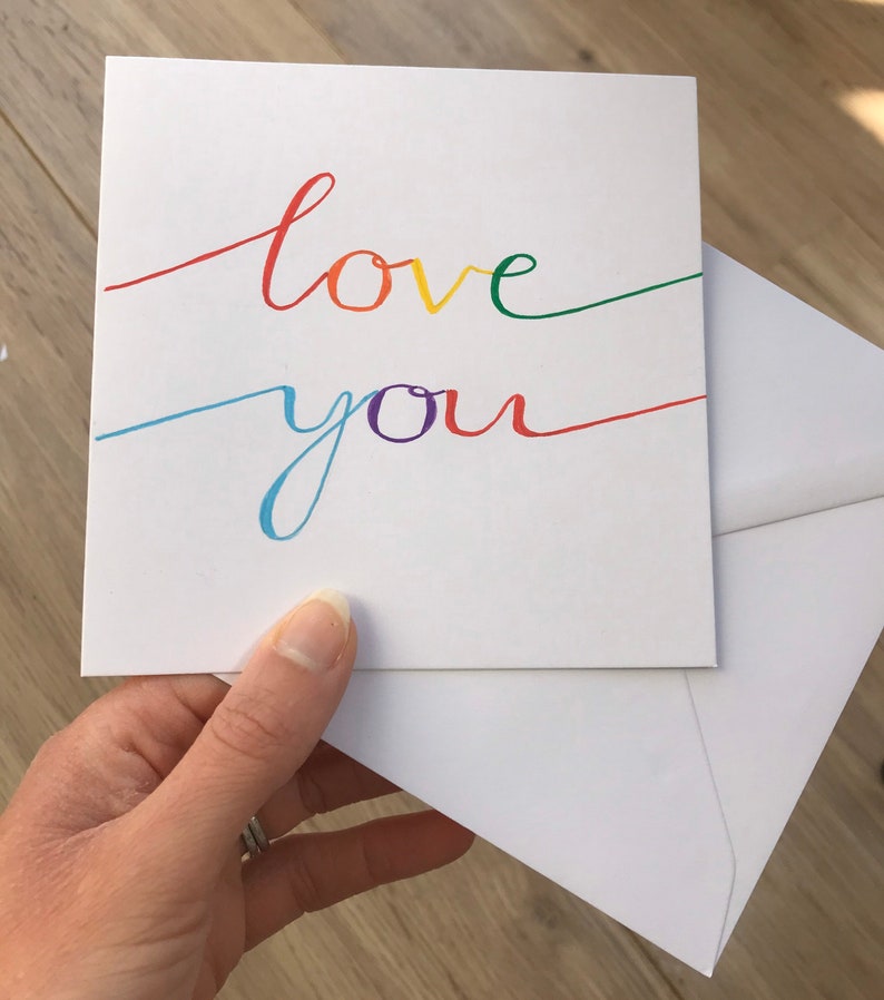 May include: A white card with the words "love you" written in rainbow colors. The card is folded in half and held in a hand.