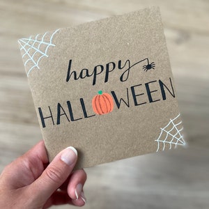 May include: A square, kraft paper Halloween card with the words "happy HALLOWEEN" in black script and block letters. An orange pumpkin is between the words. White spiderwebs are in the corners, and a black spider is above the word "Halloween".