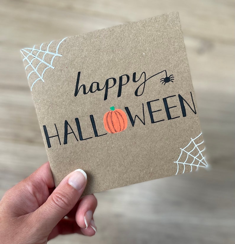 May include: A square, kraft paper Halloween card with the words "happy HALLOWEEN" in black script and block letters. An orange pumpkin is between the words. White spiderwebs are in the corners, and a black spider is above the word "Halloween".