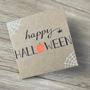 May include: A square, kraft paper card with the words "happy HALLOWEEN" in black script and block letters. A small orange pumpkin graphic is between the words. White spiderwebs are in the corners, and a black spider is above the word "HALLOWEEN".