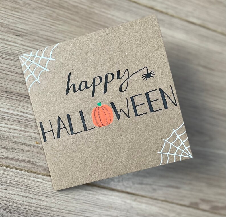 May include: A square, kraft paper card with the words "happy HALLOWEEN" in black script and block letters. A small orange pumpkin graphic is between the words. White spiderwebs are in the corners, and a black spider is above the word "HALLOWEEN".