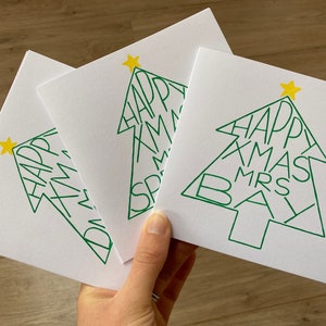 May include: Three white Christmas cards with a green tree design. The tree shape contains the words "HAPPY XMAS MRS BAY" in green. Each card has a yellow star at the top of the tree.