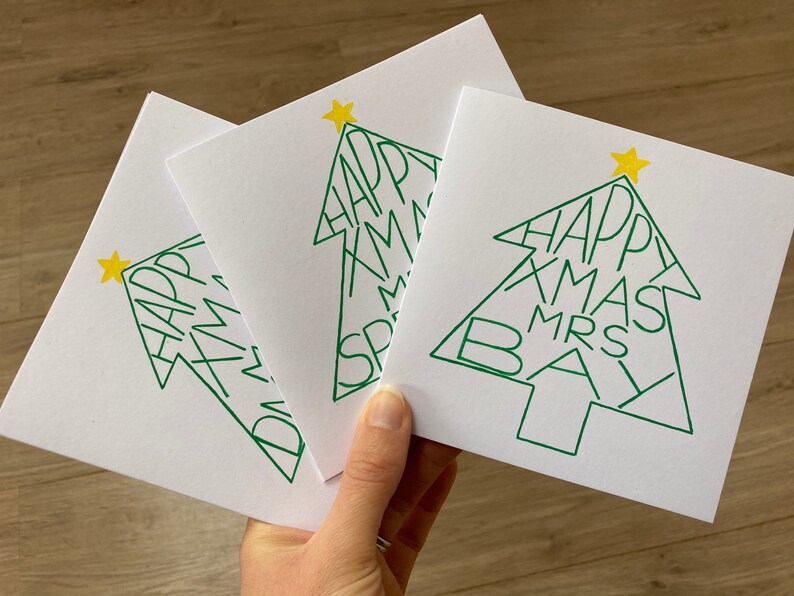 May include: Three white Christmas cards with a green tree design. The tree shape contains the words "HAPPY XMAS MRS BAY" in green. Each card has a yellow star at the top of the tree.