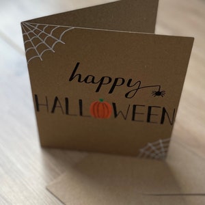 May include: A brown Halloween greeting card with the words "happy HALLOWEEN" in black script. A small orange pumpkin is centered below the word "happy". White spiderwebs are in the corners, and a small black spider is next to the word "happy".
