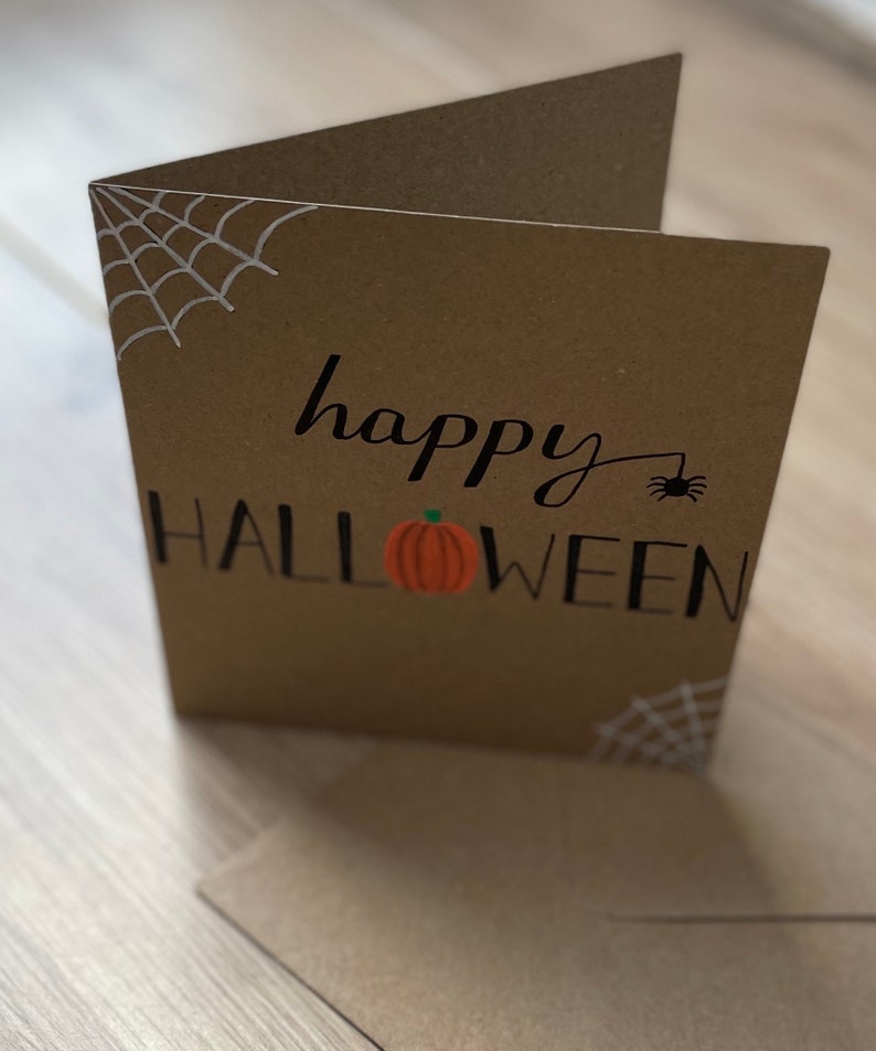 May include: A brown Halloween greeting card with the words "happy HALLOWEEN" in black script. A small orange pumpkin is centered below the word "happy". White spiderwebs are in the corners, and a small black spider is next to the word "happy".
