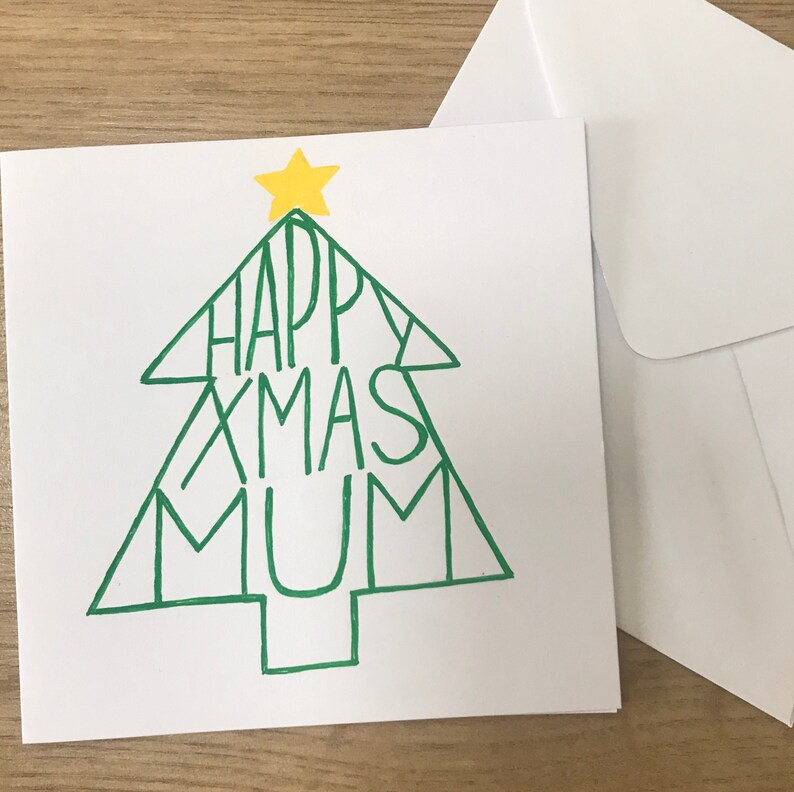 May include: A white Christmas card with a green Christmas tree design. The words "HAPPY XMAS MUM" are written inside the tree. A yellow star is at the top of the tree. A white envelope is next to the card.